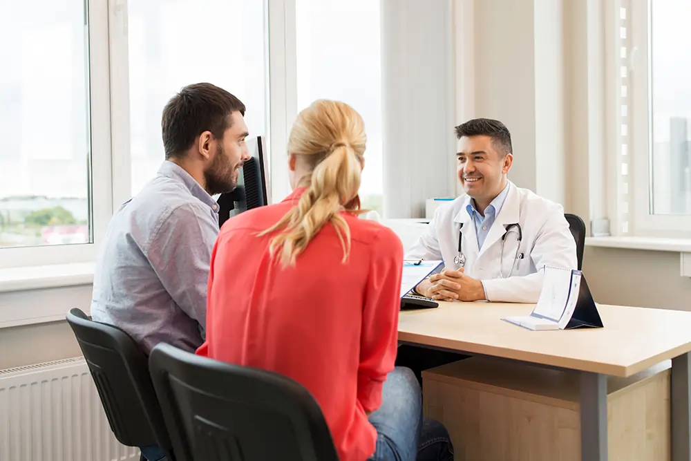 A smiling male doctor speaks with a young couple sitting across from him at his office desk.