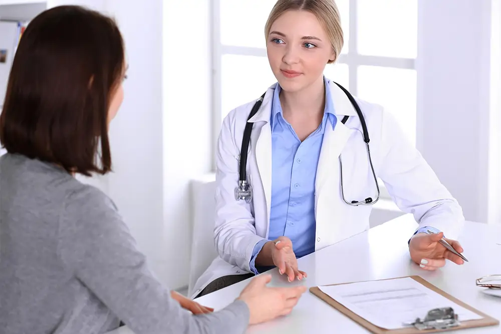 A young female doctor in a white coat is consulting with a woman at her desk.