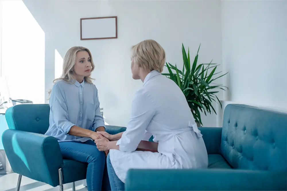 A woman is sitting on a sofa and holding hands with a professional healthcare provider.