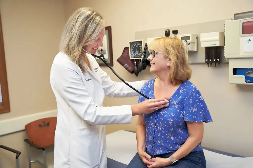 A female doctor smiles while using a stethoscope to listen to the heart of her smiling female patient during a routine checkup in an examination room.