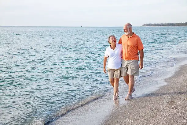 A smiling senior couple walking on a sandy beach next to the ocean.